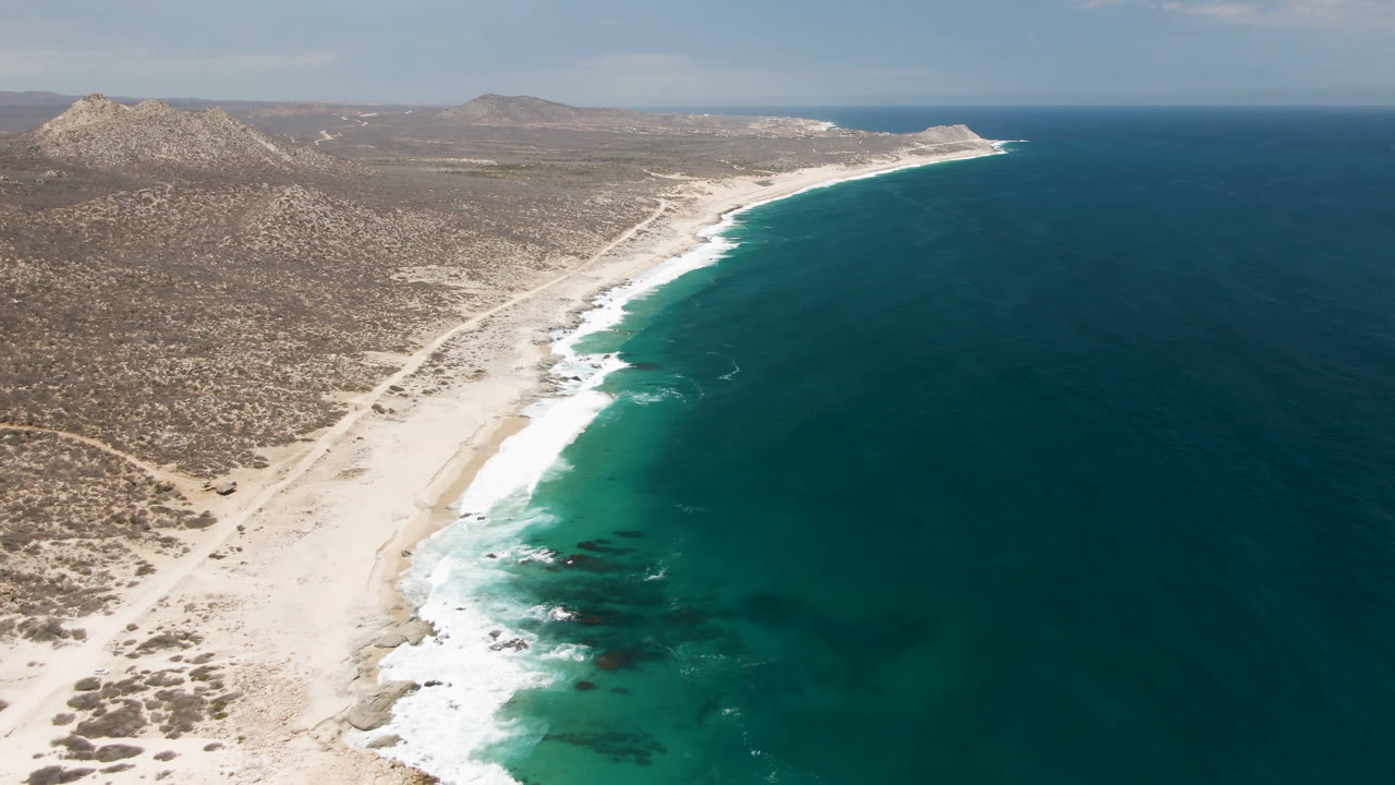 Drone-style coastline view with curved beach, surf, ocean, and desert land.