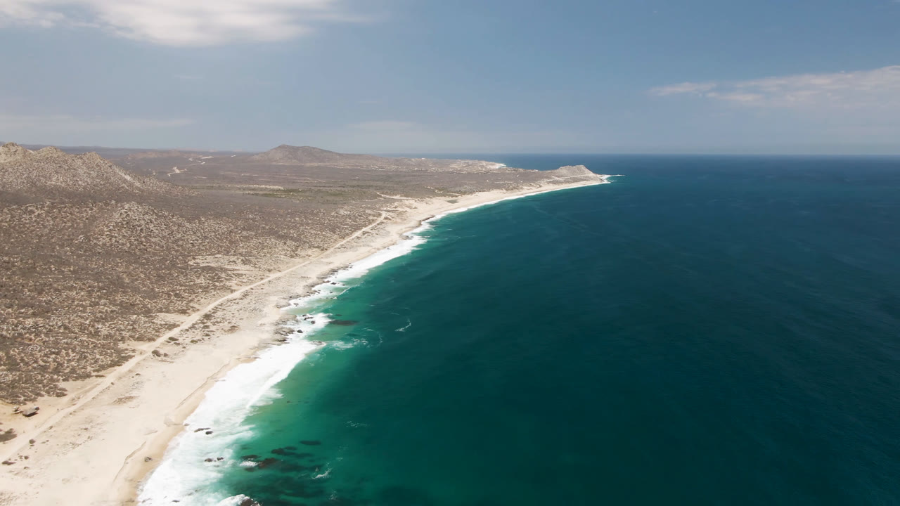 Aerial frame over Laguna Beach, Baja California Sur, with sandy shore, white surf, scrub hills, deep blue water, and no visible people.