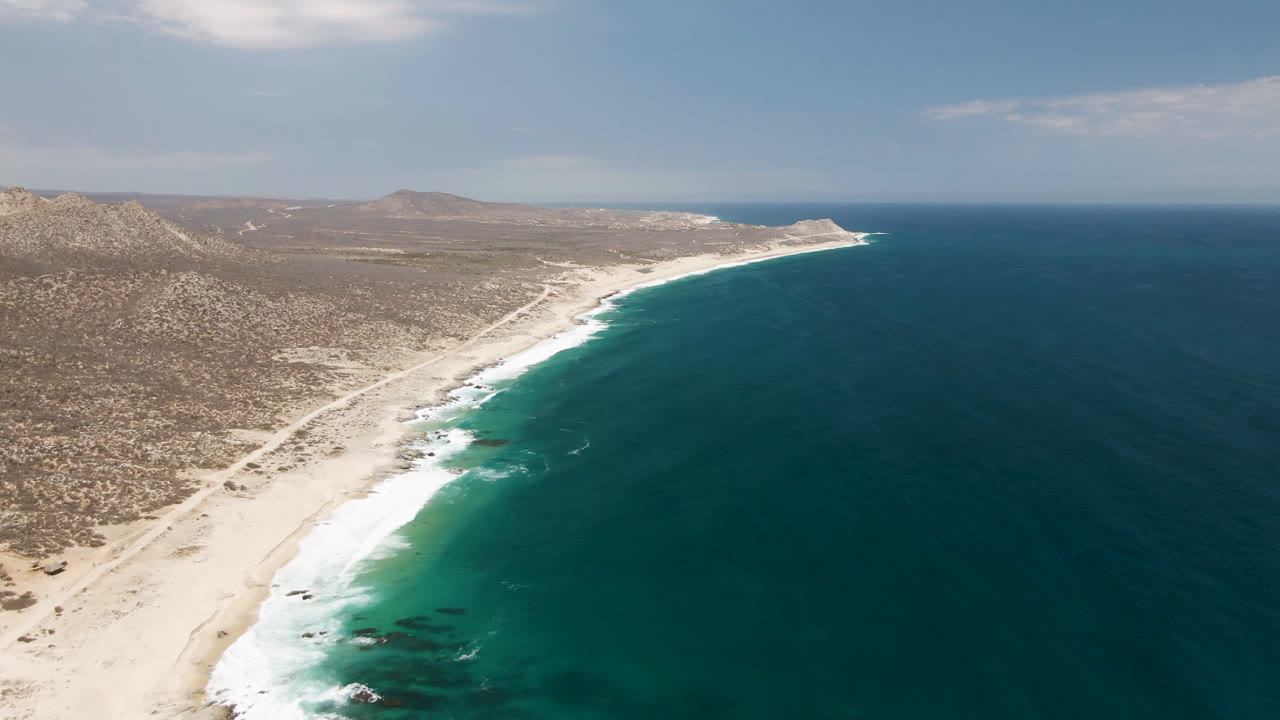 Aerial view of Laguna Beach in Baja California Sur with a curved pale beach, foamy waves, arid hills inland, and no visible people.