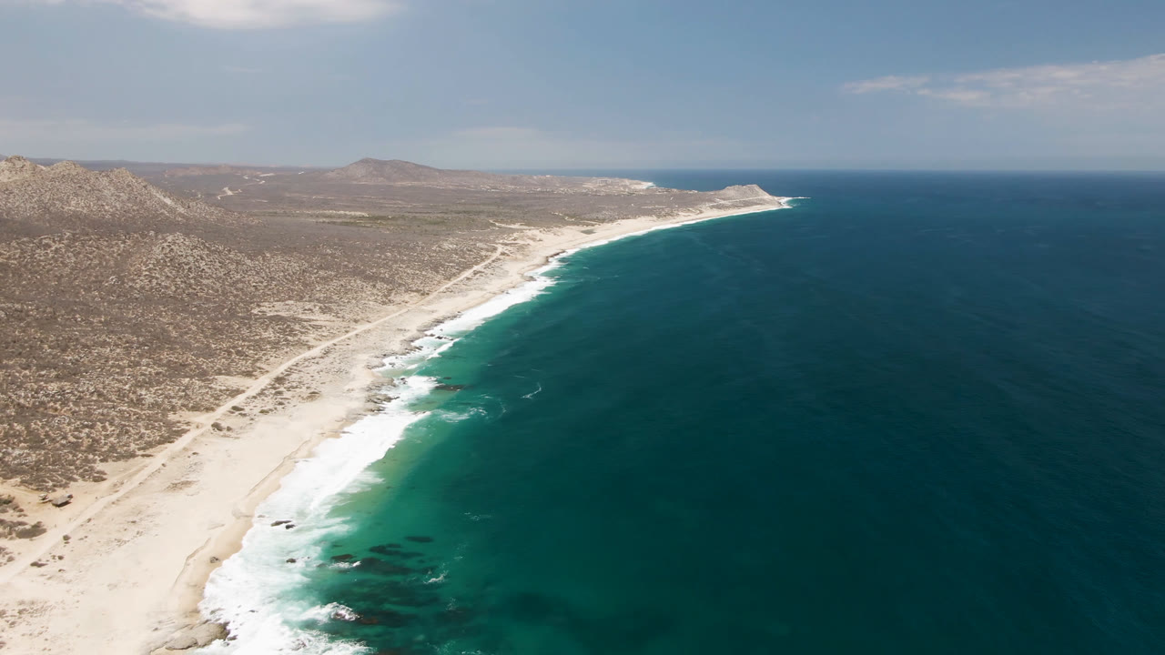 Drone-height frame over Laguna Beach, Baja California Sur, showing turquoise water near shore, darker ocean offshore, a pale beach, and no visible people.