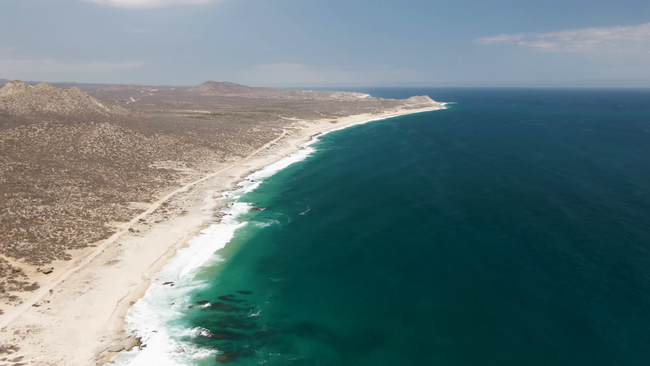 Wide aerial coastline with sandy beach, surf, hills, and open water.