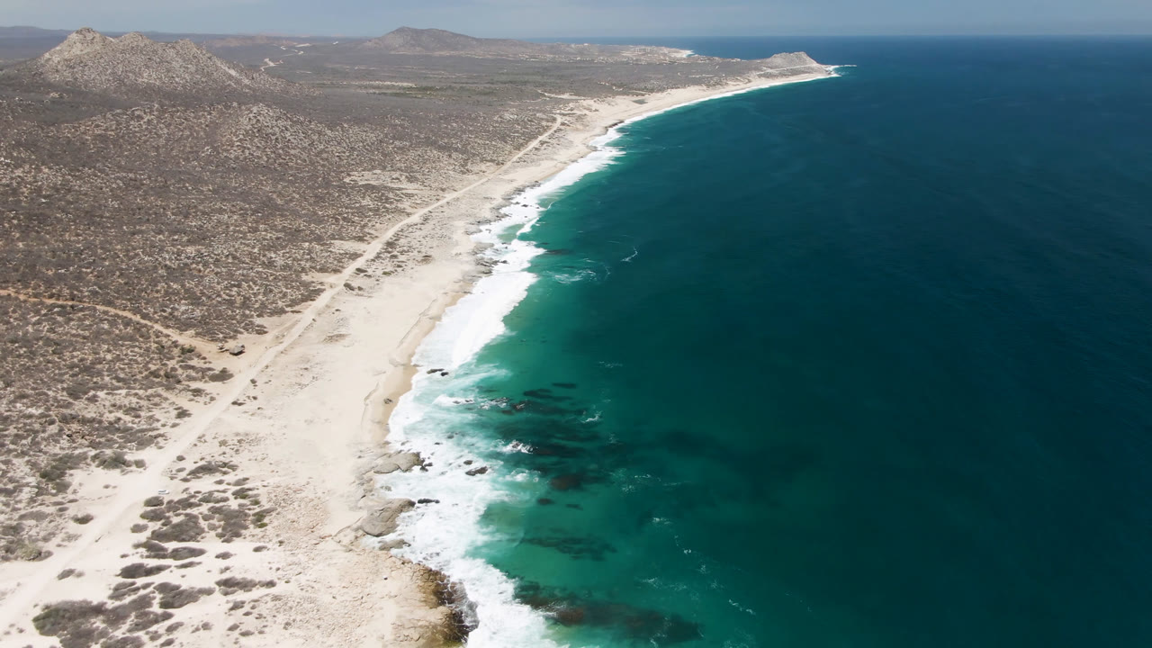 Aerial view of pale beach, turquoise surf, desert scrub, and rocky hills.