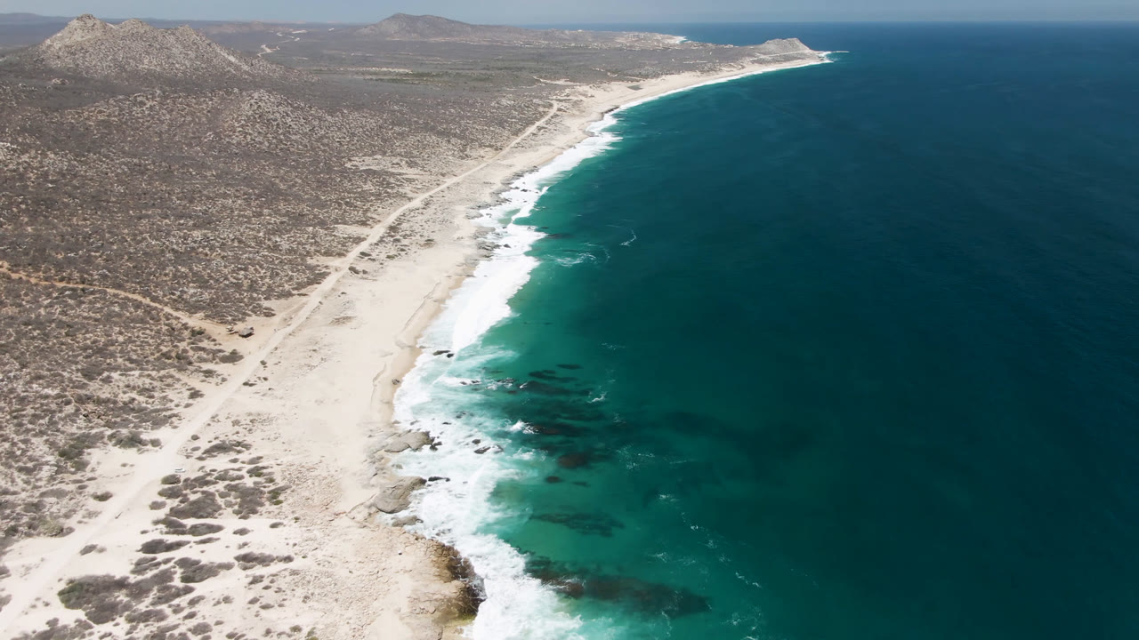 At 0:01, the Laguna Beach shoreline fills the frame with white surf bands along the sand, deeper blue water offshore, inland dirt tracks, and no visible people.