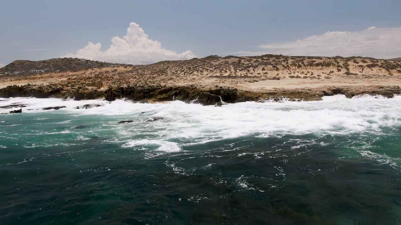White surf rolls across dark coastal rocks at Laguna Beach, Baja California Sur, with turquoise water in front, sandy scrub hills behind, and no visible people.
