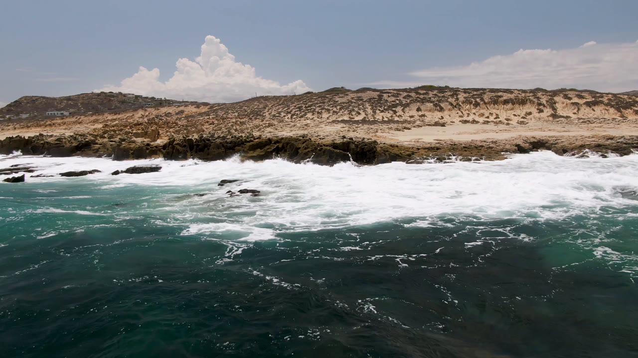Foam spreads wider over the rock shelf at Laguna Beach, Baja California Sur, with teal water in the foreground and pale dunes under a blue sky, no visible people.