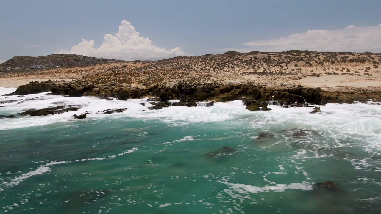 Aerial view of turquoise surf breaking over dark rocks beside a sandy beach.