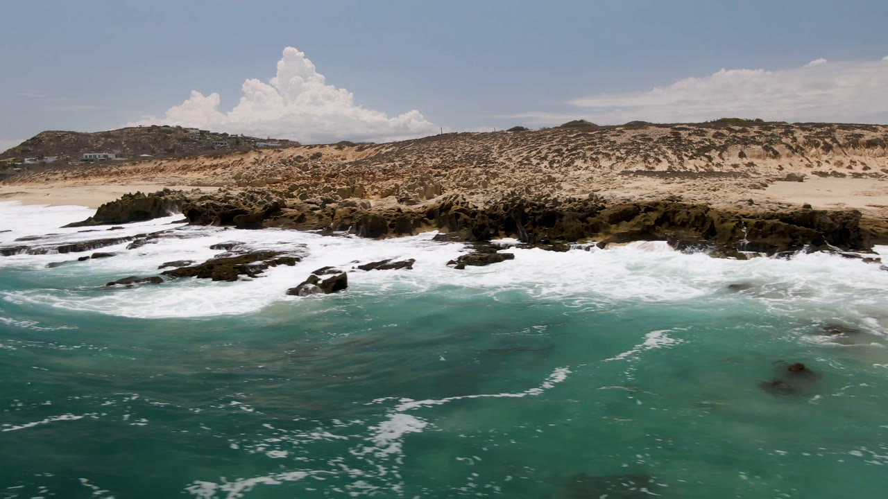At 0:01, whitewater spreads wider across the rocky shoreline at Laguna Beach, Baja California Sur, with pale sand, scrub dunes, distant low hills, and no visible people.