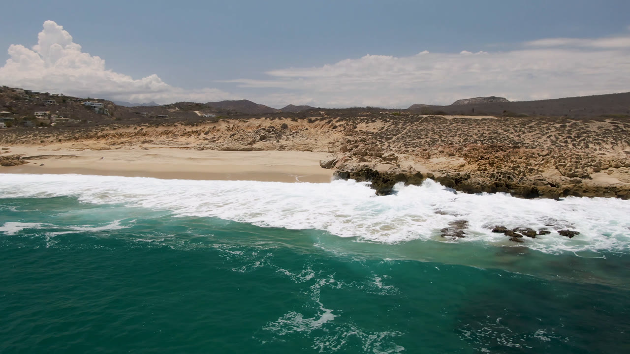 Aerial frame from Laguna Beach, Baja California Sur showing teal water in the foreground, white surf on sand and rocks, scrub-covered hills, scattered clouds, and no visible people.