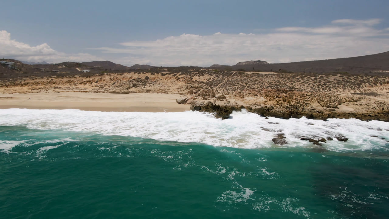 Laguna Beach aerial frame with broad white foam crossing the sand, darker rocks at the waterline, low desert vegetation, distant mountains, scattered clouds, and no visible people.