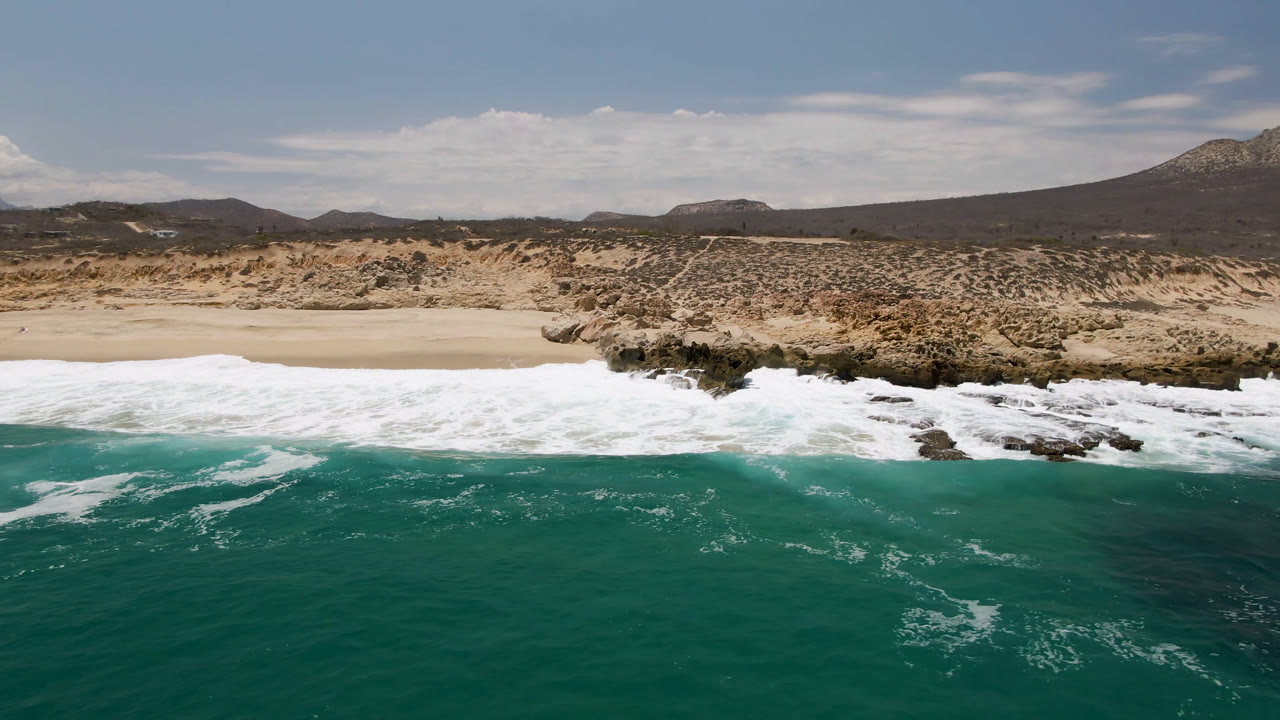 Aerial coastline with breaking waves, pale sand, rocky edges, and mountains under clouds.