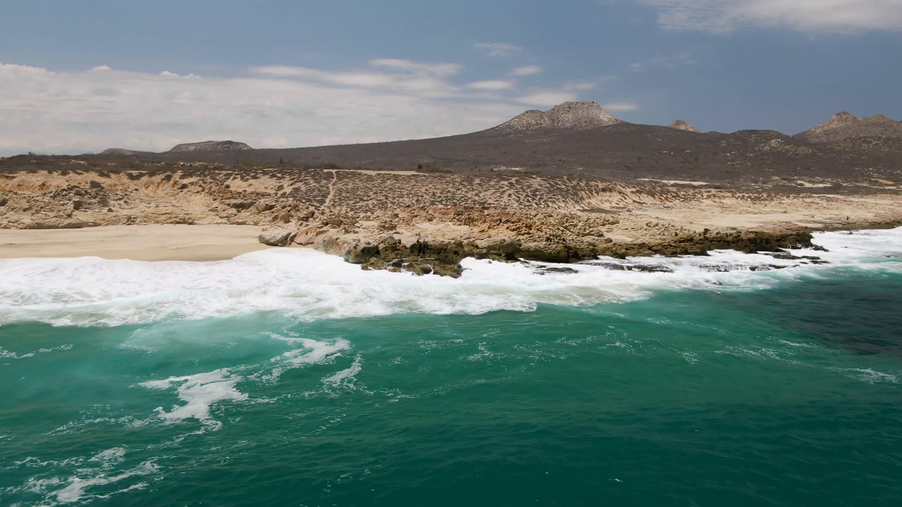 Wide aerial view of turquoise surf, sandy beach, rocky shore, and desert hills.