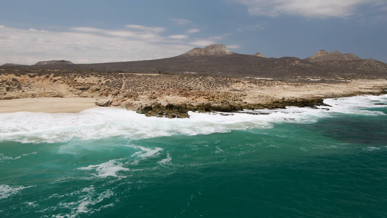 Laguna Beach, Baja California Sur coastline at 0:01 with foamy water spread across the foreground, pale beach and rocks midframe, dry scrubland, distant hills, and no visible people.