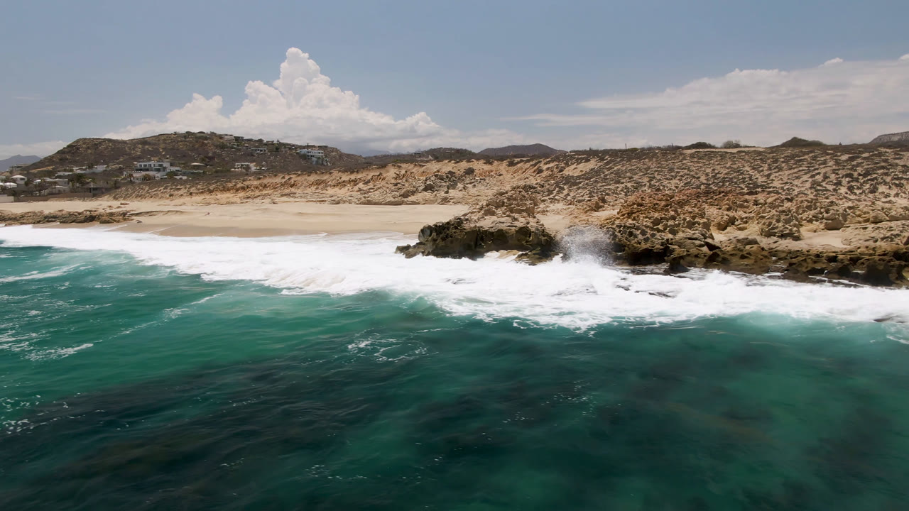 At Laguna Beach in Baja California Sur, turquoise water rolls toward a sandy beach and dark rocks beneath low scrub hills, with no visible people in this frame.