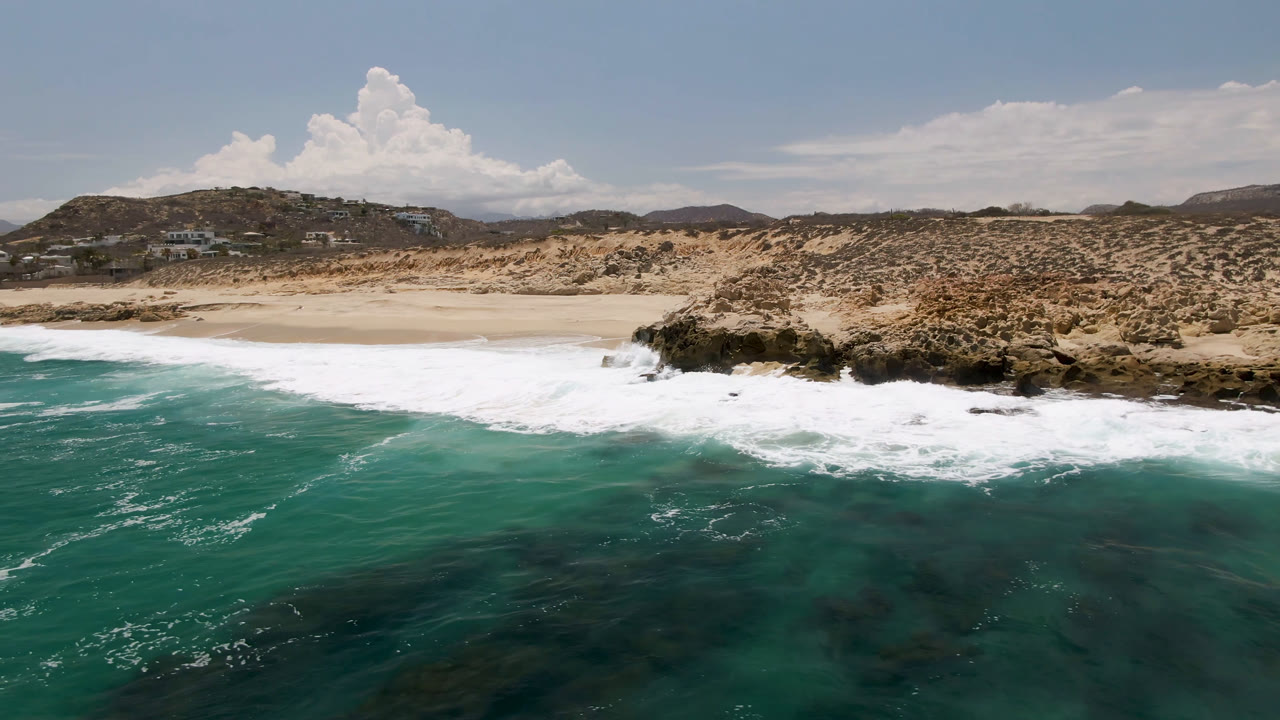 White foam spreads across the beach at Laguna Beach, Baja California Sur, with teal water in front and low brown hills behind, with no visible people in this frame.