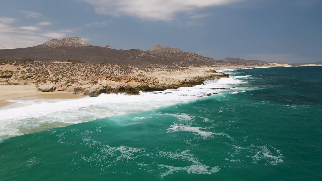 Aerial view of Laguna Beach in Baja California Sur, Mexico, with turquoise water, white surf, sandy beach, rocky desert shoreline, distant mountains, and no visible people.