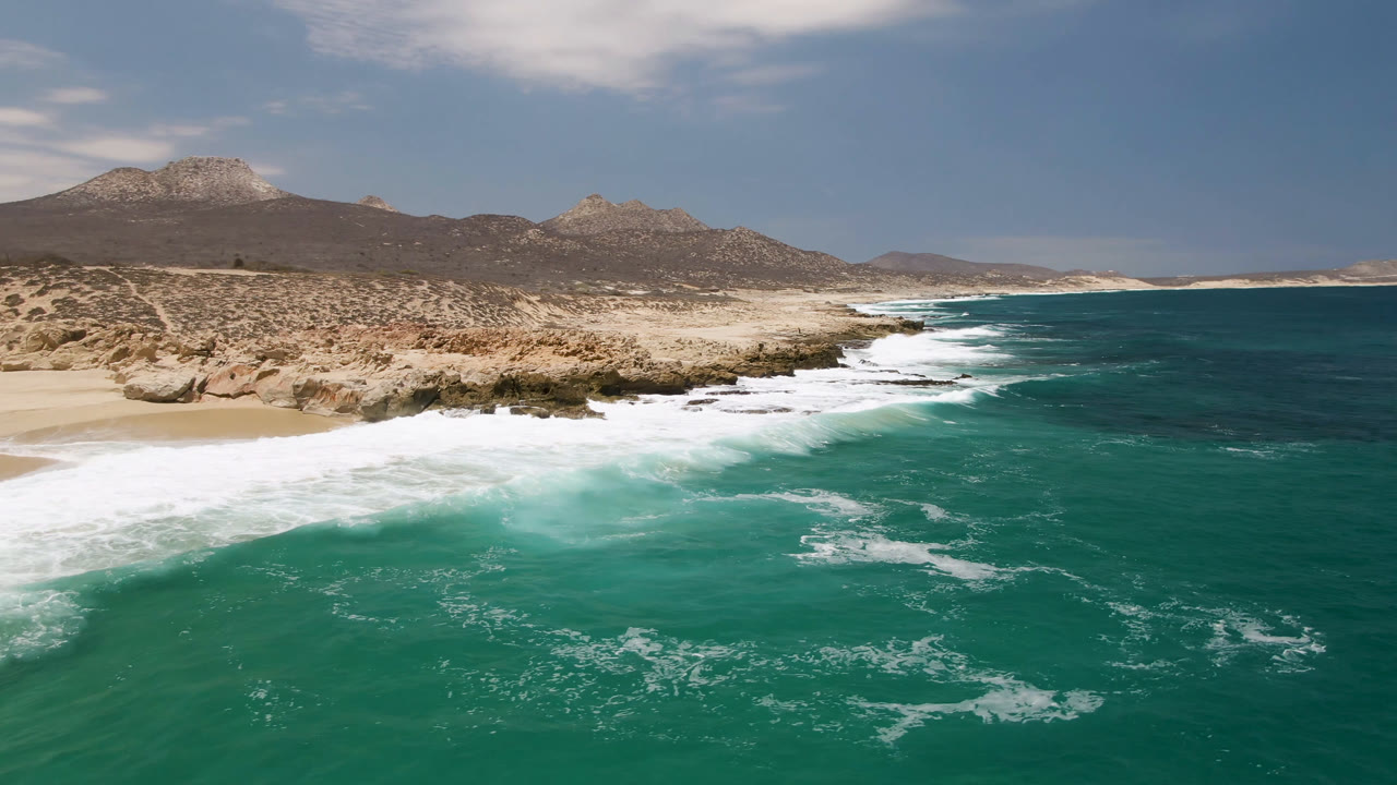 Laguna Beach shoreline in Baja California Sur, Mexico, with white waves curling toward sand, rocky desert ground to the left, mountains in the distance, and no visible people.