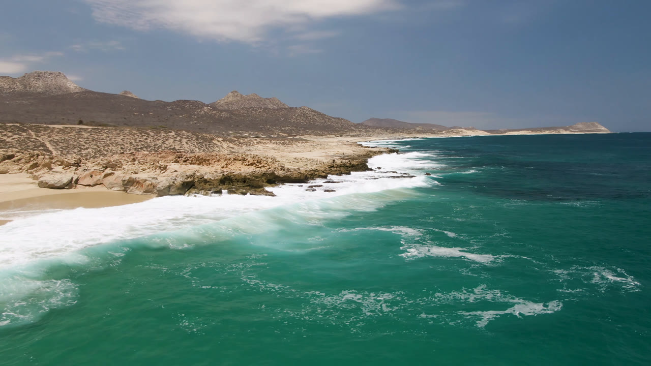 Turquoise ocean spreads across the foreground below a sandy beach and mountain-backed coast.