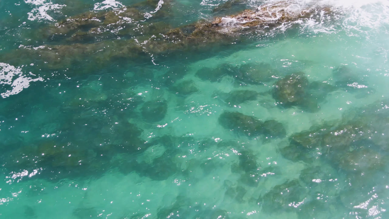 Overhead view of turquoise water and darker submerged rocks at Laguna Beach, Baja California Sur, Mexico, with white foam entering the top of the frame and no visible people.