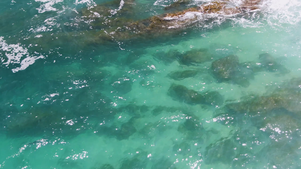 Aerial frame of clear teal water over dark reef patches at Laguna Beach, Baja California Sur, Mexico, with small white flecks of foam and no visible people.