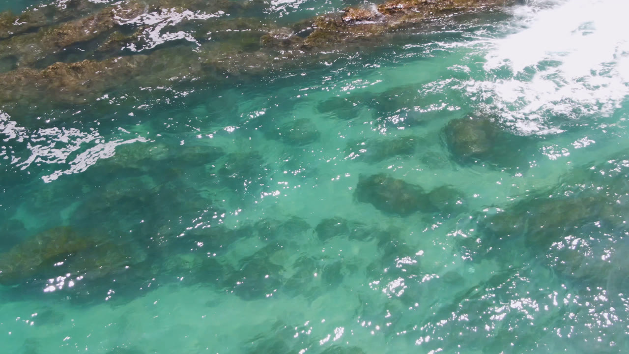 Overhead view of turquoise water at Laguna Beach, Baja California Sur, with dark submerged rocks, a band of white surf foam near the top, and no visible people.