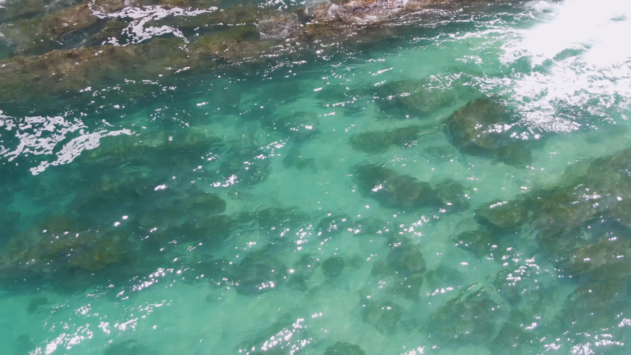 Aerial frame over Laguna Beach, Baja California Sur, showing clear green-blue water, scattered dark rocks below the surface, small white ripples, and no visible people.