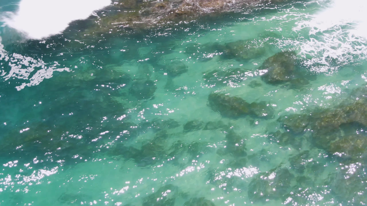 Overhead Laguna Beach water frame with turquoise shallows, darker reef patches under the surface, white breaking foam across the upper edge, and no visible people.