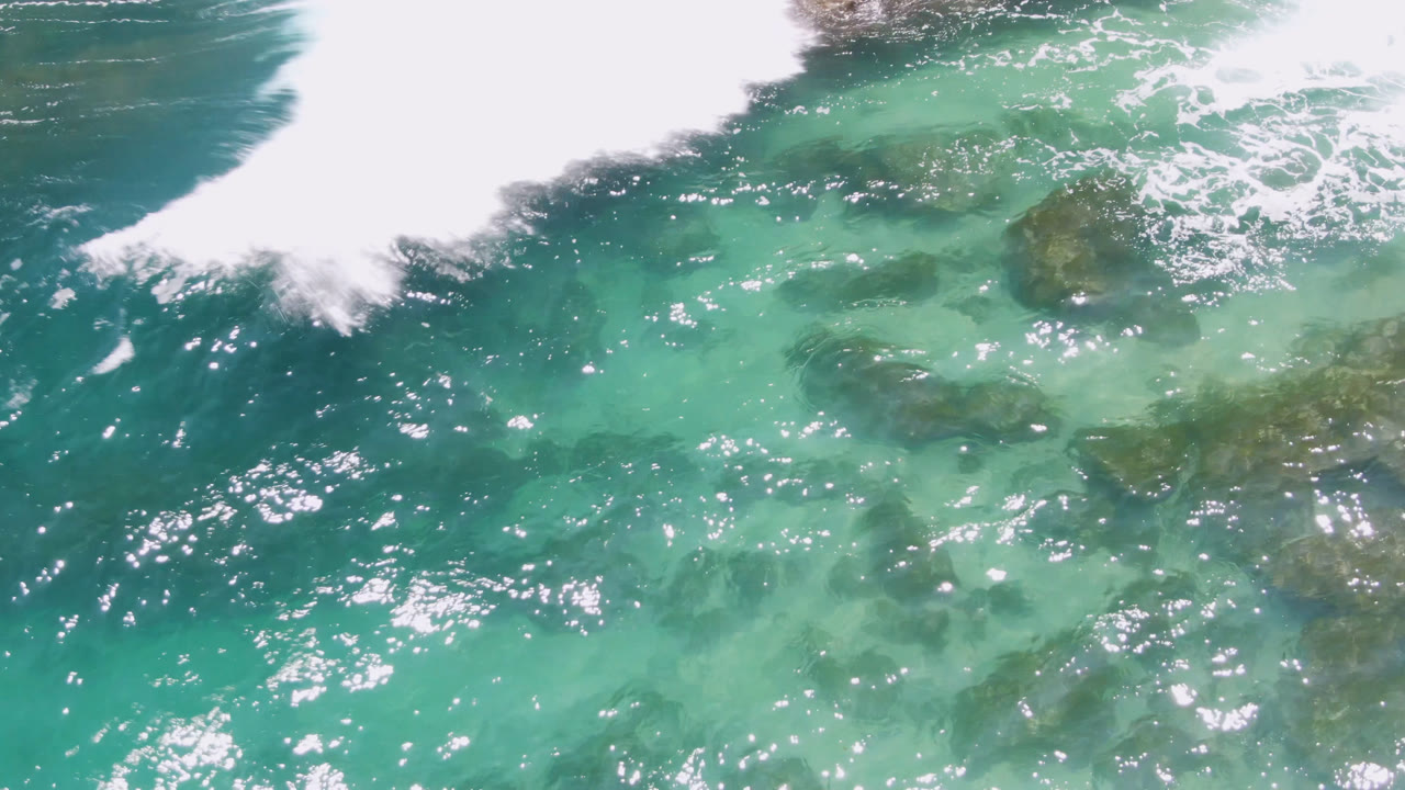 Aerial view at Laguna Beach, Baja California Sur, where pale turquoise water covers dark rock patches and white foam spreads in from the upper left, with no visible people.
