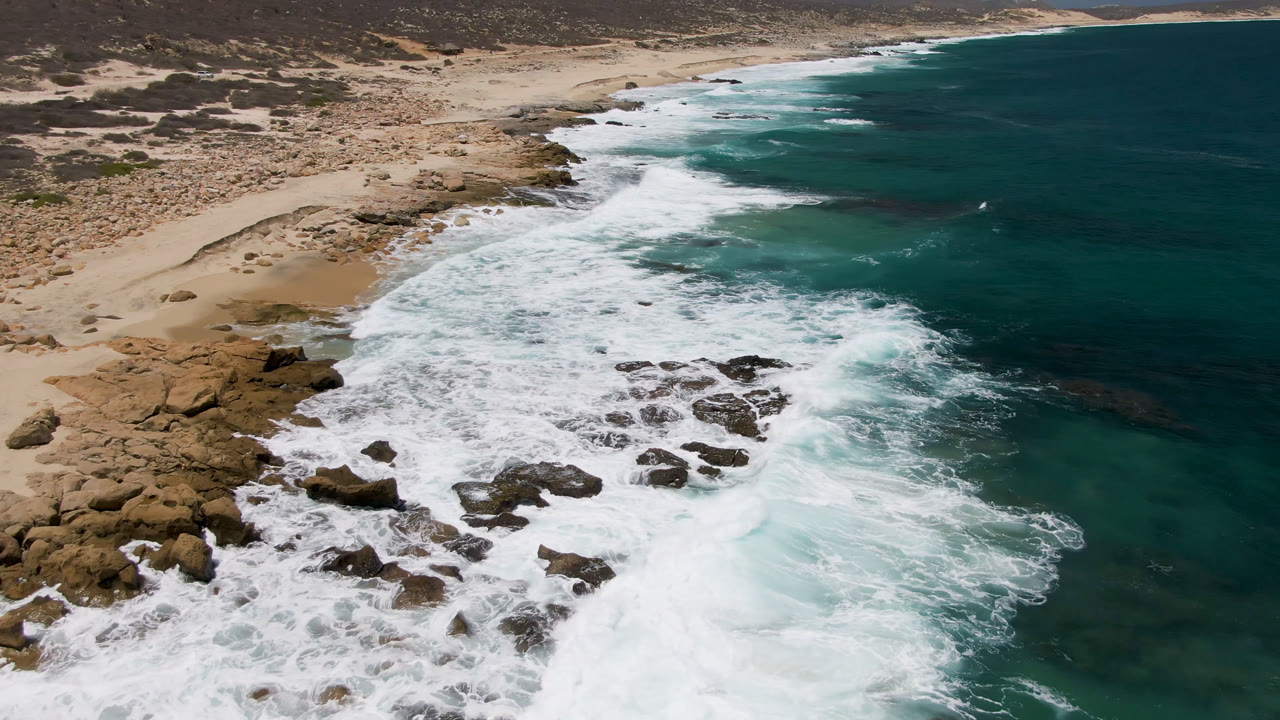 Aerial frame of Laguna Beach, Baja California Sur, with white surf spilling over dark shoreline rocks, pale sand at left, teal ocean at right, dry scrubland beyond, and no visible people.