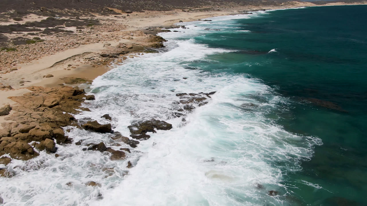 Aerial view of Laguna Beach, Baja California Sur, where a broader sheet of white foam covers the rocks near shore, with sandy beach and scrubland behind and no visible people.