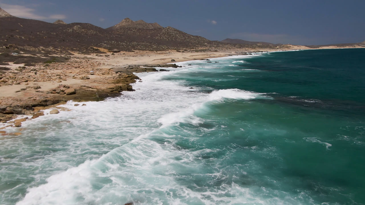 Aerial view of Laguna Beach in Baja California Sur with white surf washing toward rocks and sand, turquoise ocean to the right, mountains in the distance, and no visible people.