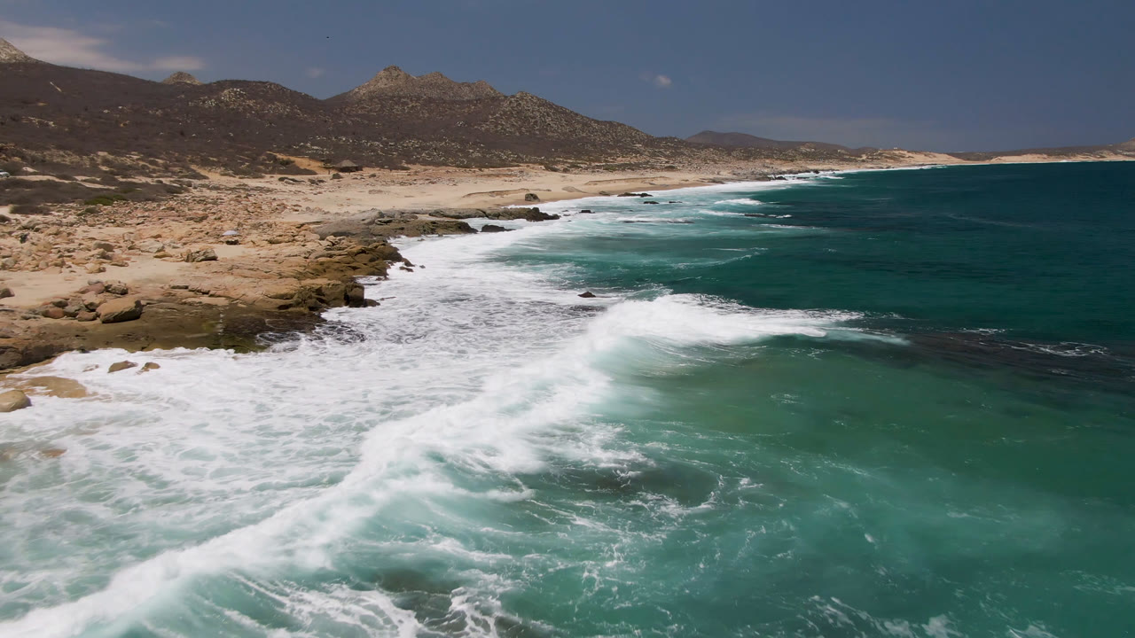 Laguna Beach aerial frame with a broad band of seafoam crossing shallow turquoise water, tan sand and rocks on the left, distant mountain ridges, and no visible people.