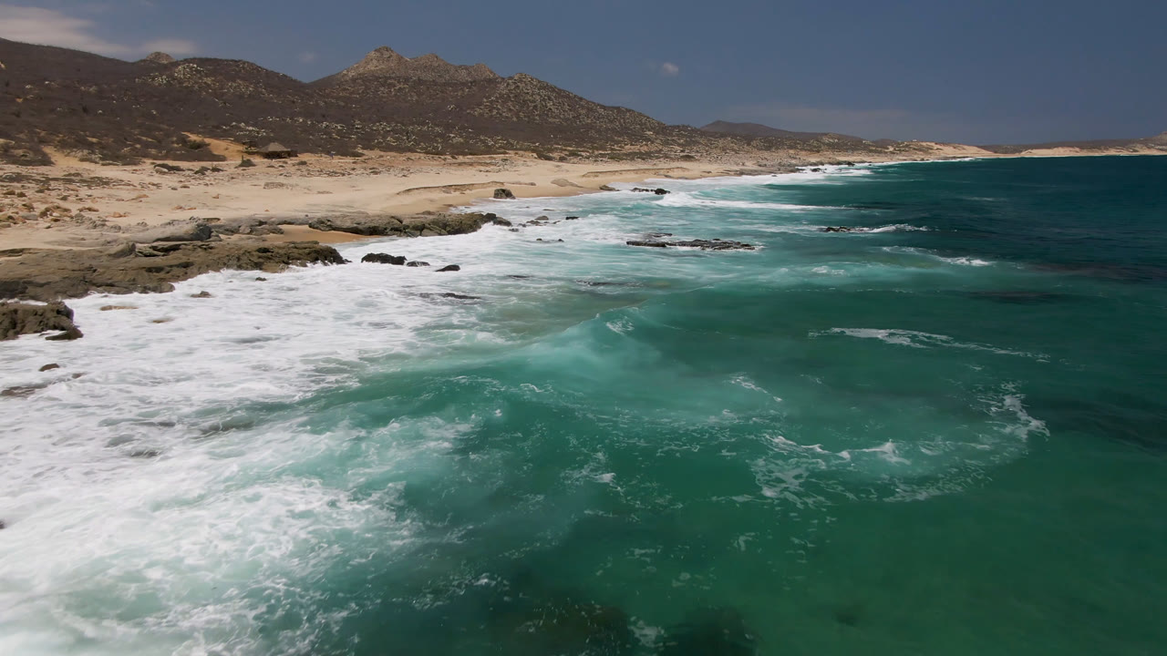 Aerial view of Laguna Beach in Baja California Sur, Mexico, with turquoise water, white surf, sandy shore, rocky outcrops, low hills, and no visible people.