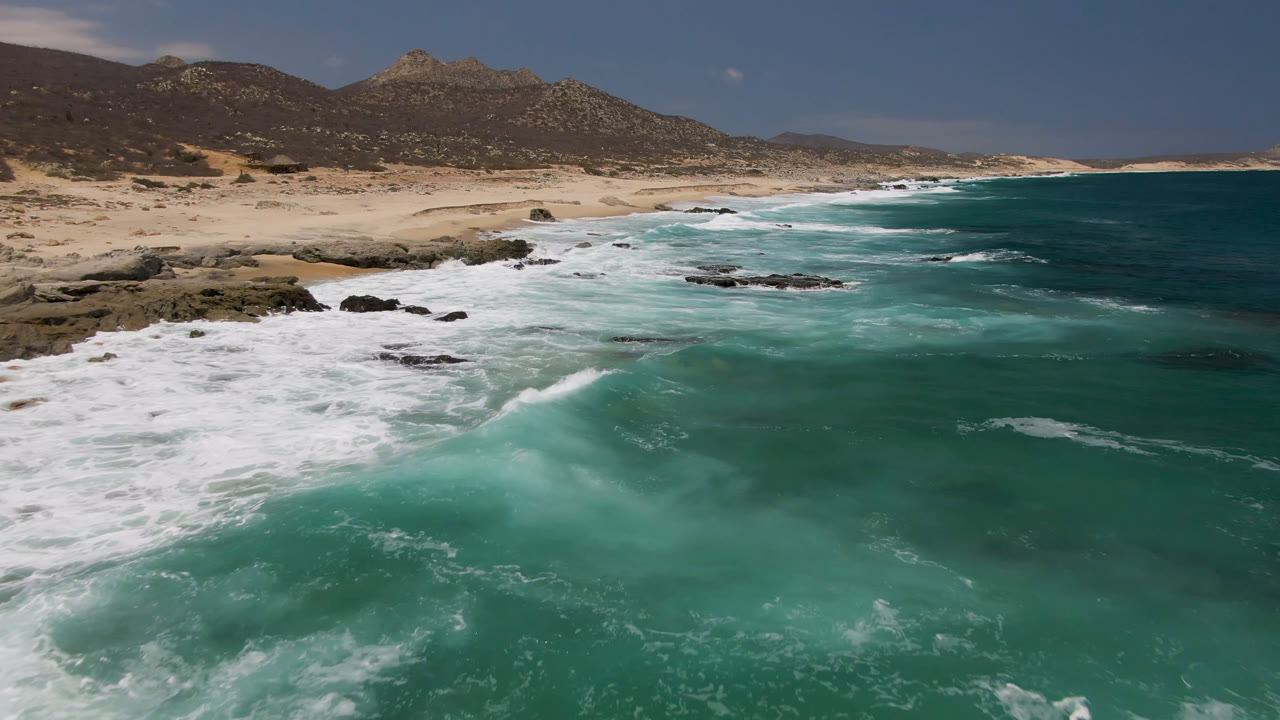 Elevated view of Laguna Beach, Baja California Sur, as white foam spreads across teal water near a pale sand beach and dark shoreline rocks, with no visible people.