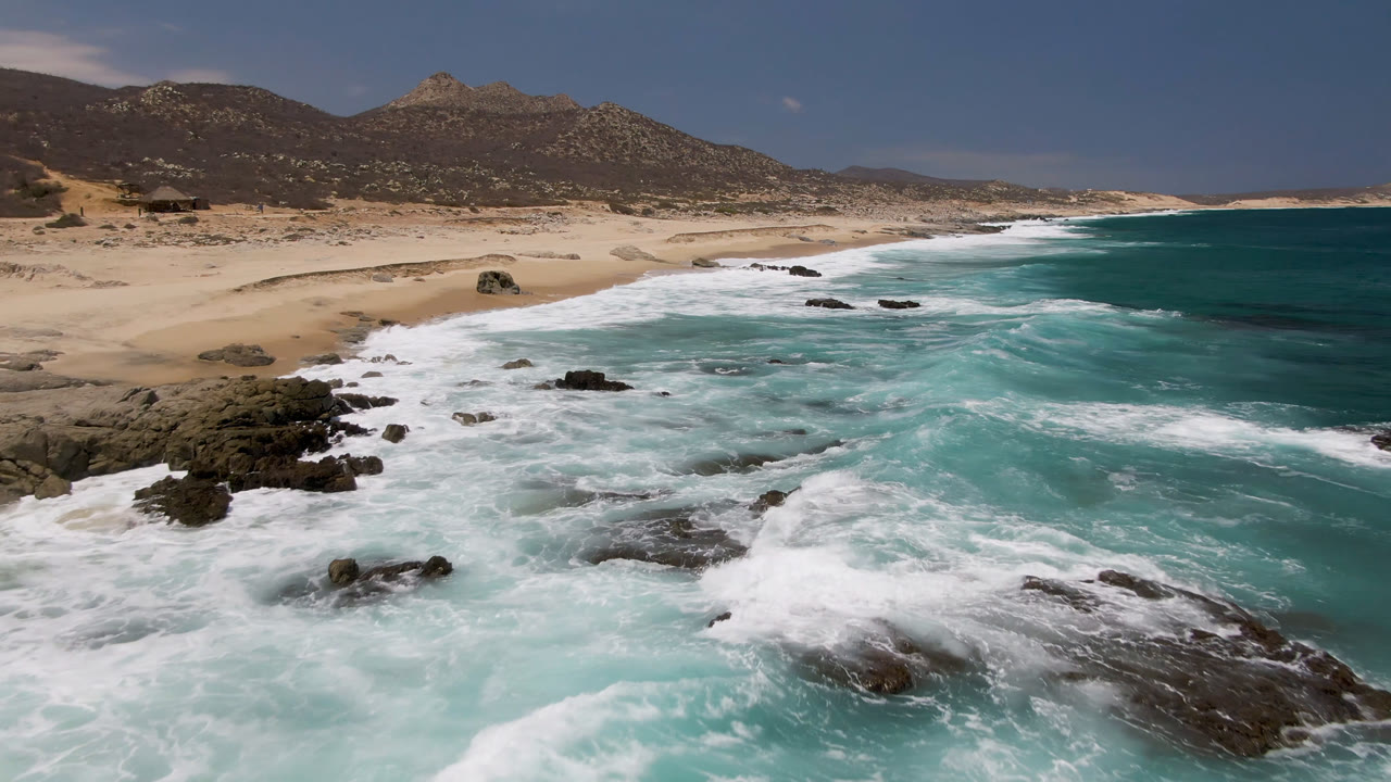 Elevated view of turquoise surf breaking over dark rocks beside a sandy beach.