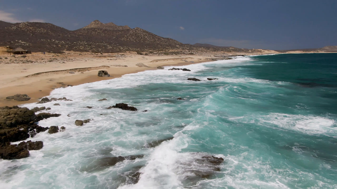 Laguna Beach, Baja California Sur, Mexico, seen from above with foam bands pushing toward the sand, scattered rocks in the water, desert hills inland, and no visible people.