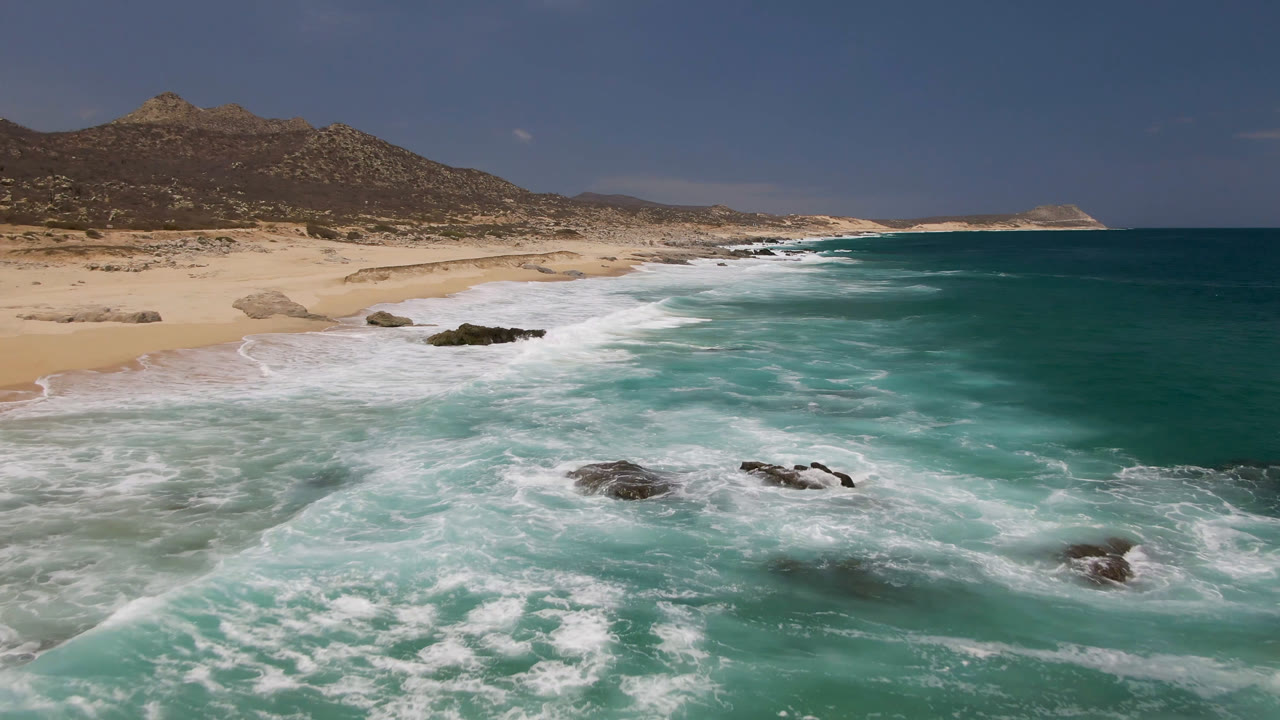 Aerial beach view with turquoise surf, sand, rocks, and mountain backdrop.