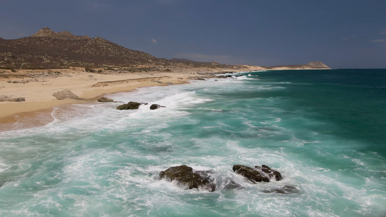 Laguna Beach coastline seen from above as white foam spreads across turquoise water beside a broad sand curve, with rocky outcrops and a mountain inland, and no visible people.