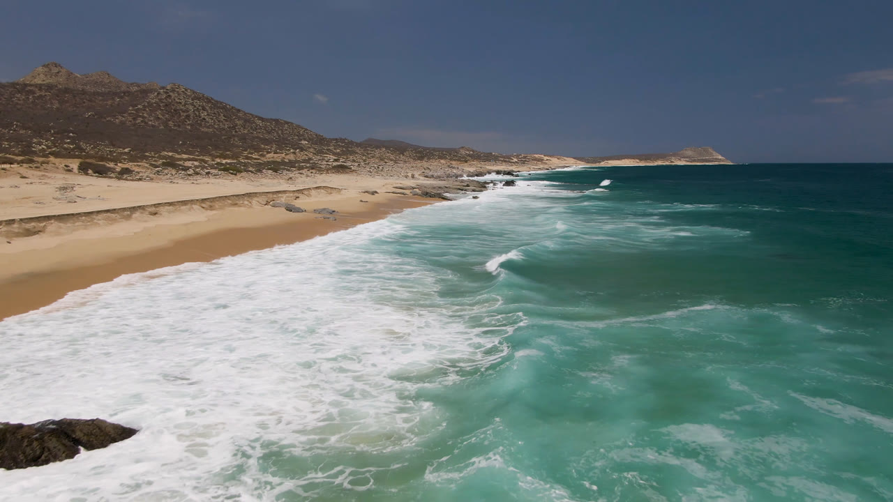 Laguna Beach, Baja California Sur shoreline with white surf entering from the lower left, turquoise water to the right, tan sand and dry hills beyond, and no visible people.