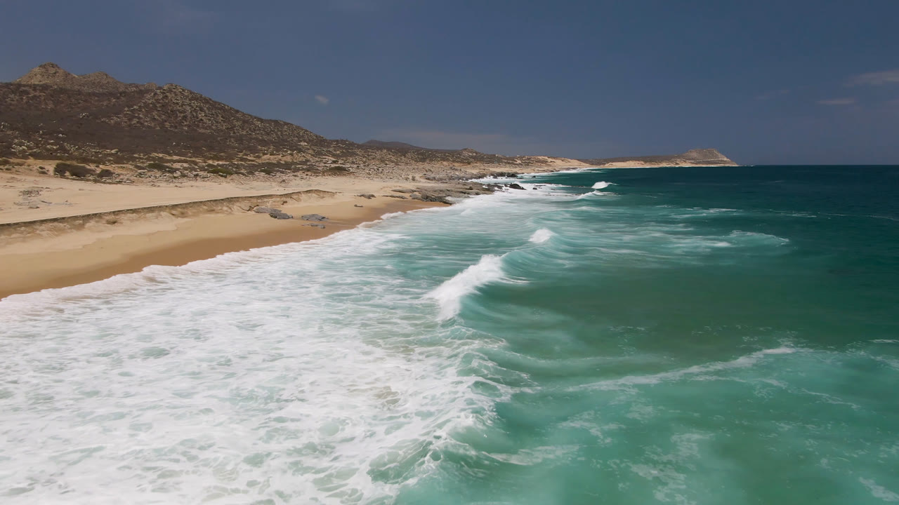 Laguna Beach, Baja California Sur coast with a broader sheet of white foam across the foreground water, open sand on the left, low rocky hills behind, and no visible people.