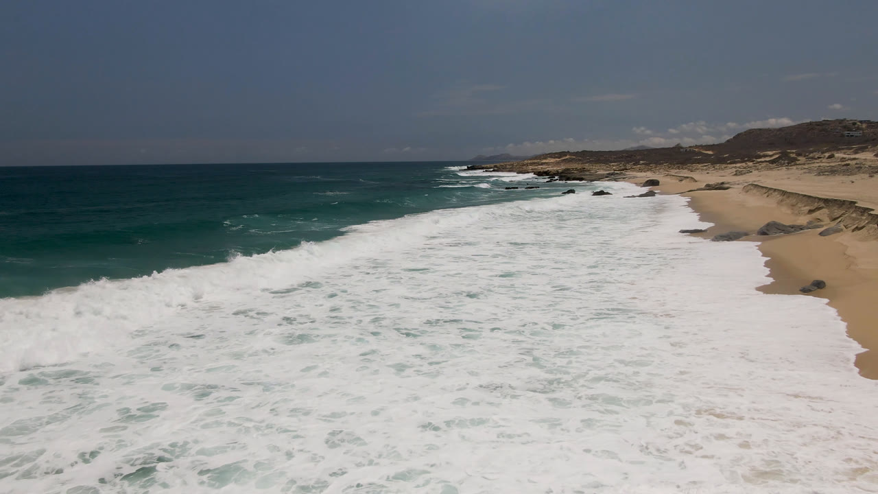 Aerial view of Laguna Beach, Baja California Sur, with broad white surf crossing the foreground, teal water at left, sandy beach at right, distant hills, and no visible people.