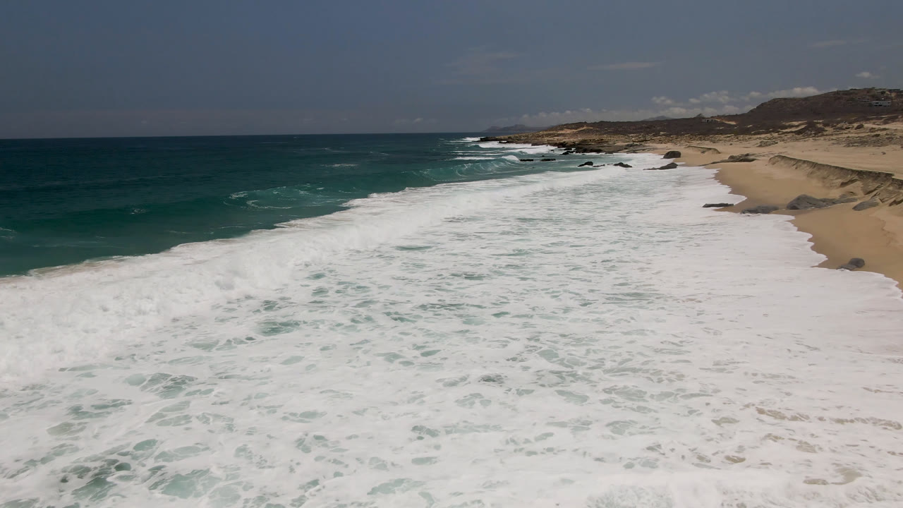 Laguna Beach surf spreads across the center of the frame, with darker ocean on the left, tan sand on the right, scattered rocks near the shore, and no visible people.