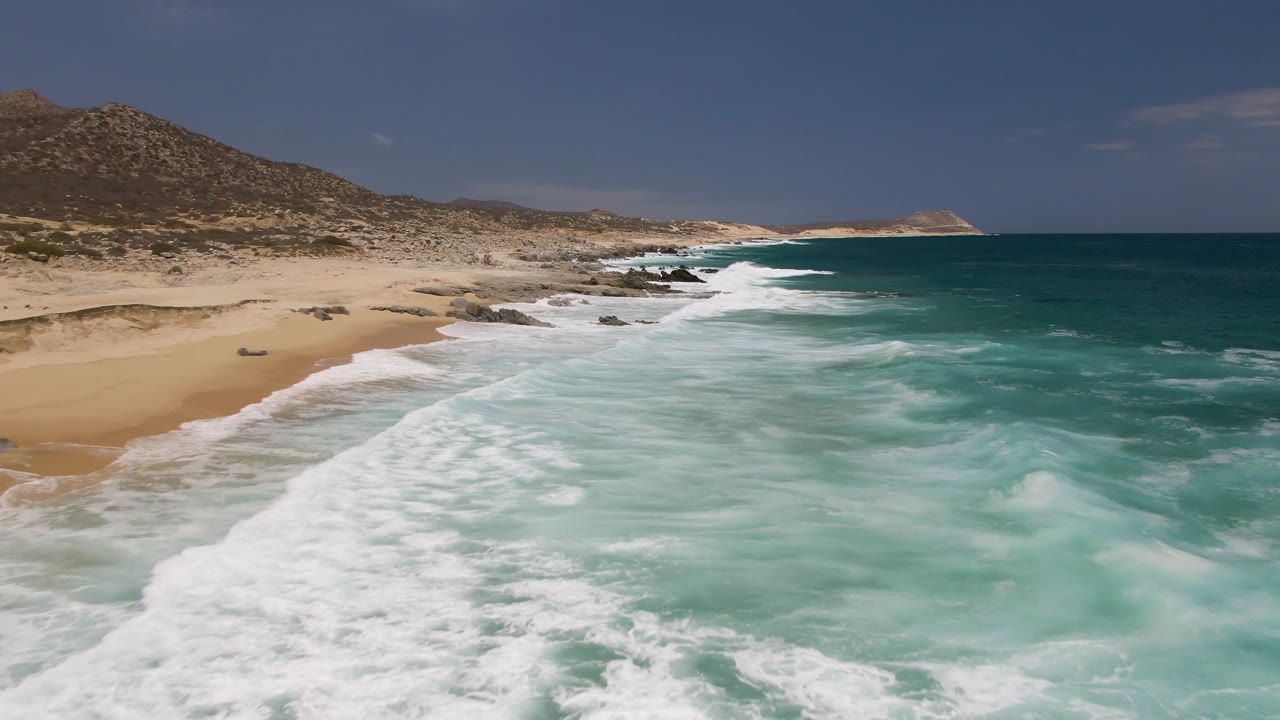 Aerial view at Laguna Beach, Baja California Sur, with white surf spreading over turquoise water beside pale sand, rocky shoreline, dry hills, and no visible people.