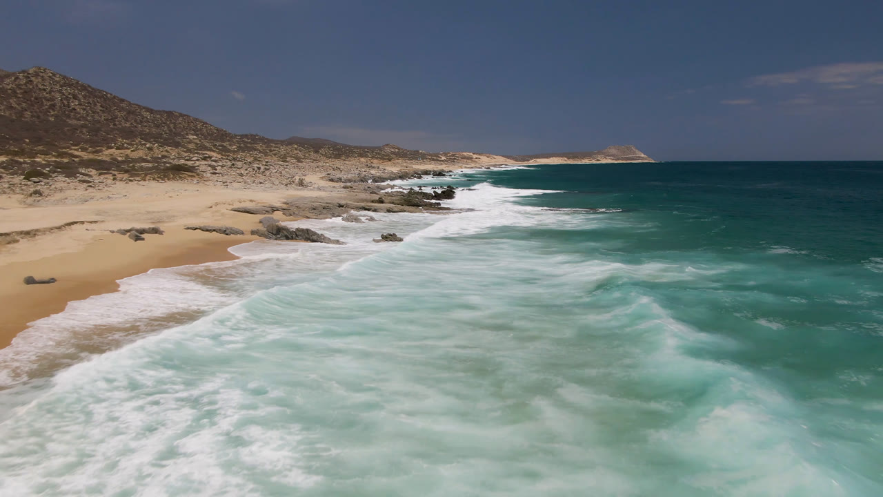 Laguna Beach, Baja California Sur shoreline with whitewater pushing diagonally along the sand, dark rocks near the break, dry hillside at left, and no visible people.
