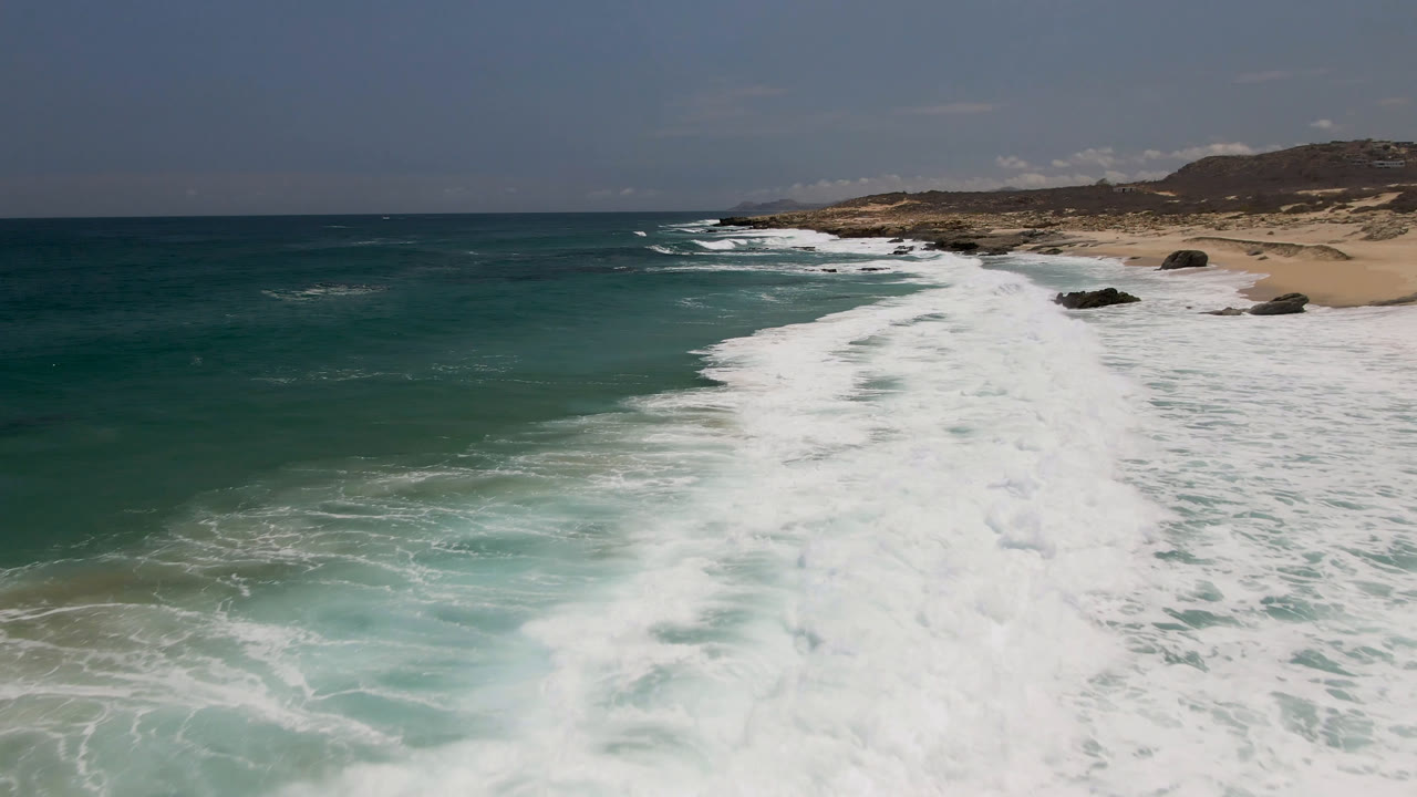 Laguna Beach aerial frame with a long band of whitewater running diagonally toward pale sand, darker offshore water on the left, scattered rocks near shore, and no visible people.
