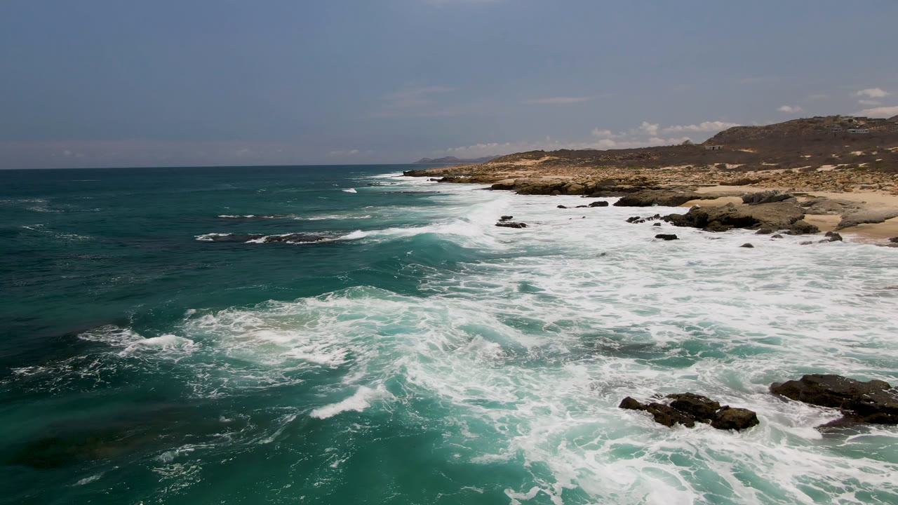 Green ocean surf spreads across dark rocks below a tan Laguna Beach, Baja California Sur shoreline, with low hills in the distance and no visible people.