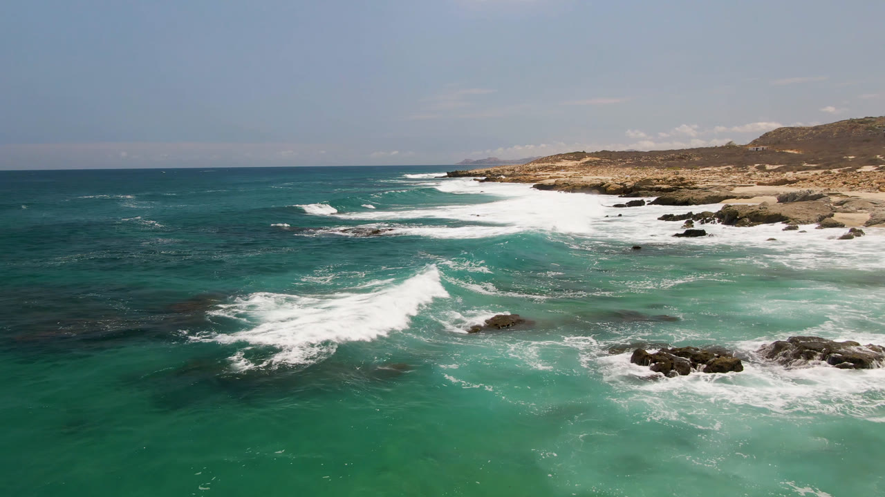 Aerial view at Laguna Beach, Baja California Sur, with turquoise water in the foreground, white surf crossing dark rocks, sandy shore beyond, and no visible people.