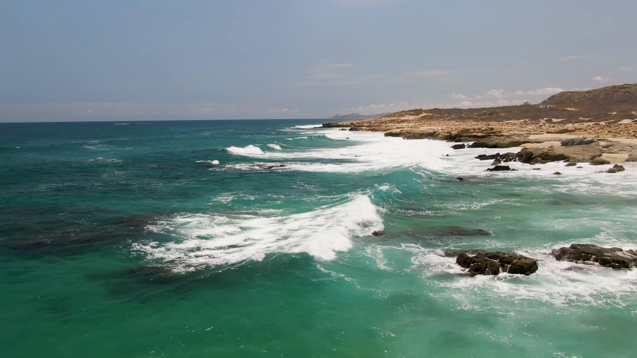 At Laguna Beach, Baja California Sur, white water spreads farther across the turquoise shallows while dark rocks sit near the lower right and no visible people appear.