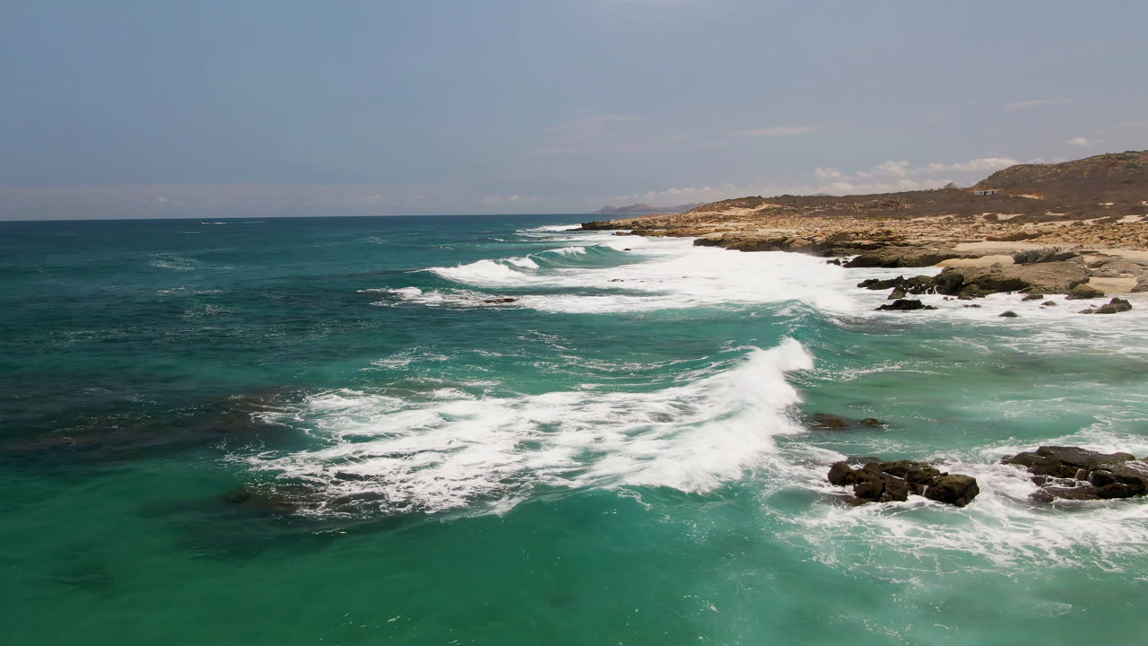 Aerial coastal view with surf foam crossing rocks and open ocean to the left.