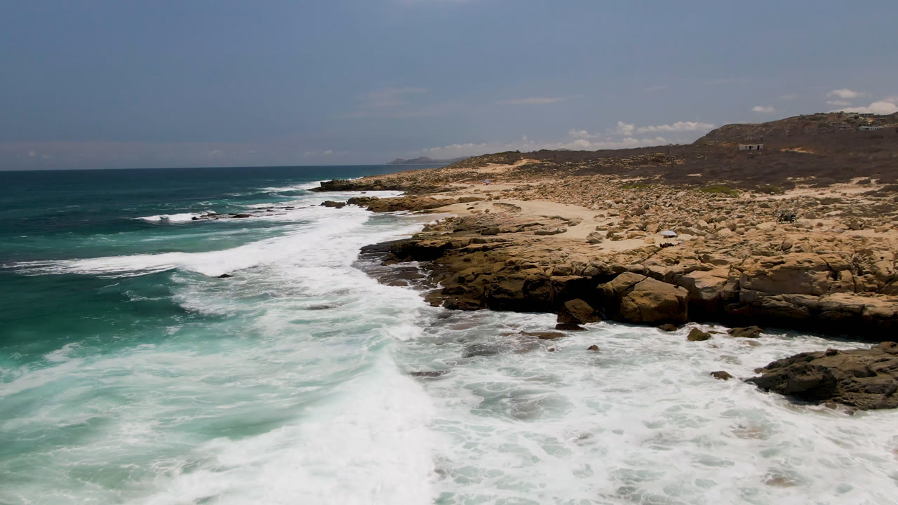 Laguna Beach, Baja California Sur coastline with white surf filling the left foreground, tan rocks and sand to the right, distant hills, and no visible people.
