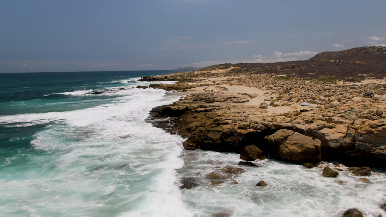 Laguna Beach, Baja California Sur shoreline with a broad white wave line crossing the midground, teal water at left, rock shelves at right, and no visible people.